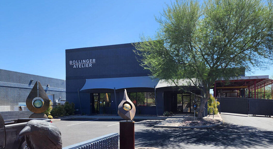 A color photo of the front of Bollinger Atelier, a dark grey building with the name at the front, next to a bright green palo verde tree with an abstract sculpture installed in the front.