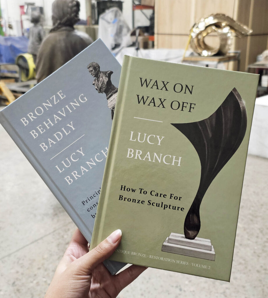 A hand holding up copies of “Wax on Wax Off” and “Bronze Behaving Badly,” two books by Lucy Branch that James has shared with the metal artisans.
