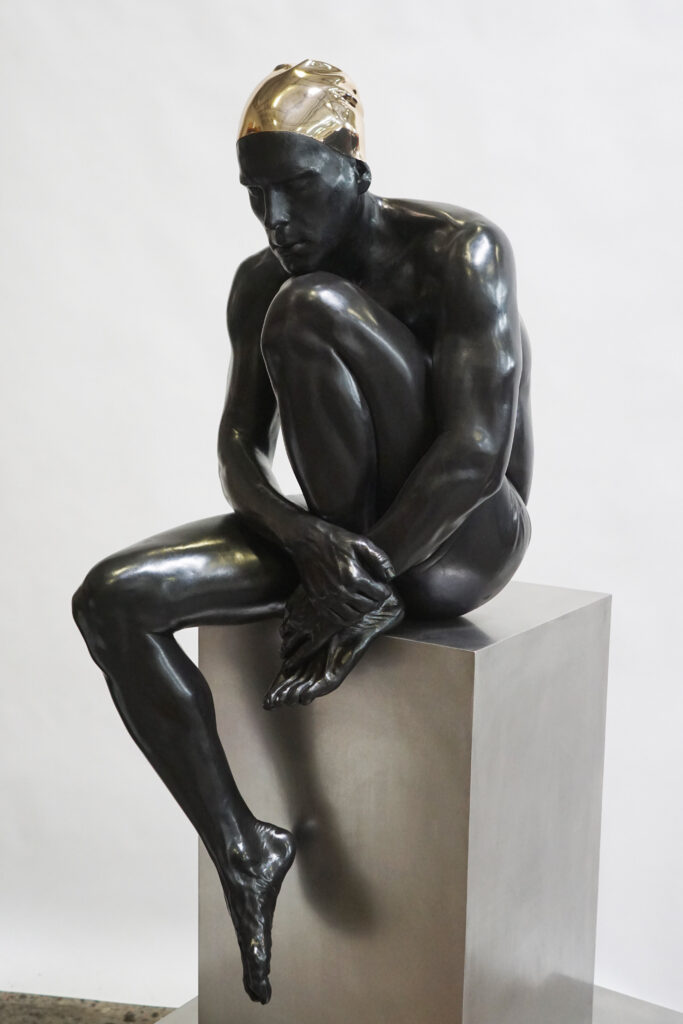 A studio photo of Carole Feuerman’s “The Thinker” - a jet black patinated sculpture of a male figure at rest with one leg up under his chin, and the other dangling off the pedestal.