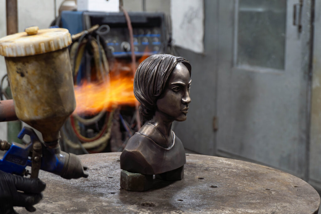The sculpture is being patinated in the metals shop. The patina artisan is spraying a chemical to deepen the brown color of the sculpture, and adding heat with a flame torch. 