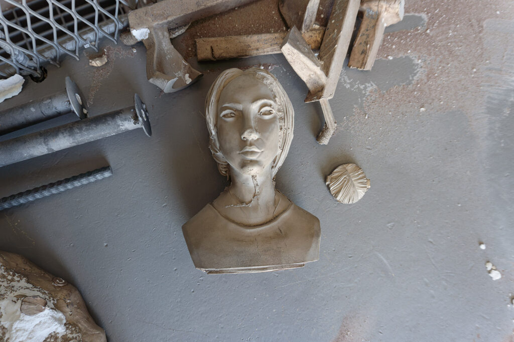 The portrait has been cast in bronze. The rough casting sits atop a metal shelf with other pieces of cast bronze, bits of removed shell, and tools.